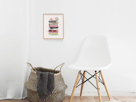 A white chair and a woven basket with a blanket sit by a white wall. Above them hangs the TS Book Stack Art Print by Onderkast Studio, perfect for Swifties or Taylor Swift fans who love colorful decor and cozy reading nooks.