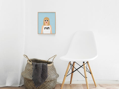 A minimalist room featuring a white chair, a woven basket with a blanket, and the Onderkast Studio TS 1989 Art Print—depicting a woman in sunglasses and a 1989 shirt—hanging on a white wall.