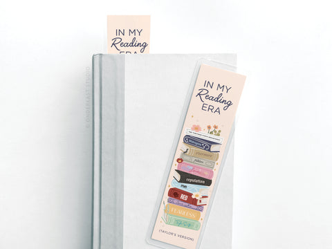 A white hardcover book displays the Onderkast Studio TS In My Reading Era Laminated Bookmark, featuring vibrant print with IN MY Reading ERA and a stack of colorful books named after Taylor Swift albums; another matching bookmark peeks out.