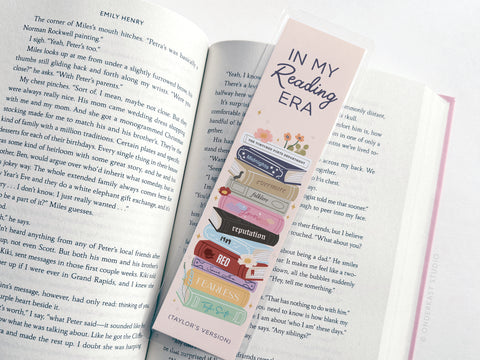 The TS In My Reading Era Laminated Bookmark by Onderkast Studio features vibrant print, an In My Reading Era message, and a stack of books titled with Taylor Swift albums like Red, Reputation, and Fearless (Taylor’s Version).