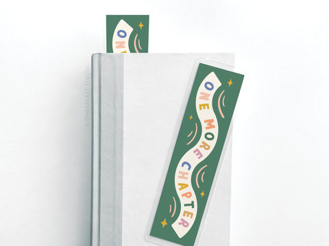 The Onderkast Studio One More Chapter Laminated Bookmark features colorful lettering on green and is ideal for avid readers, with one bookmark peeking from closed pages and another resting flat atop a white book.