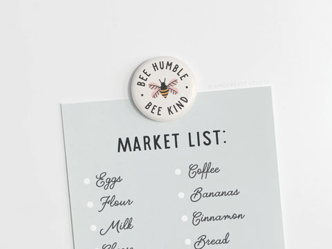 The Bee Humble Bee Kind Button Magnet by Onderkast Studio, featuring a playful bee design, holds a light blue market list on a white surface with items like eggs, flour, milk, cheese, coffee, bananas, cinnamon, and bread.