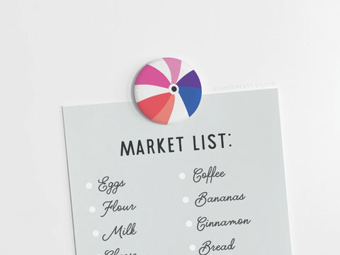 The Onderkast Studio Beachball Button Magnet keeps your market list—eggs, flour, milk, coffee, bananas, cinnamon, bread—secure on a light grey surface. Its playful beach ball design adds a pop of color and style to your space.