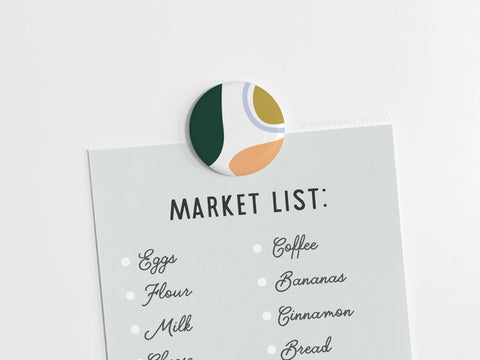 An Onderkast Studio Abstract No. 1 Button Magnet – DISCONTINUED—brightly colored and round—holds a pale blue Market List sheet with eggs, flour, milk, coffee, bananas, cinnamon, and bread written in black script.