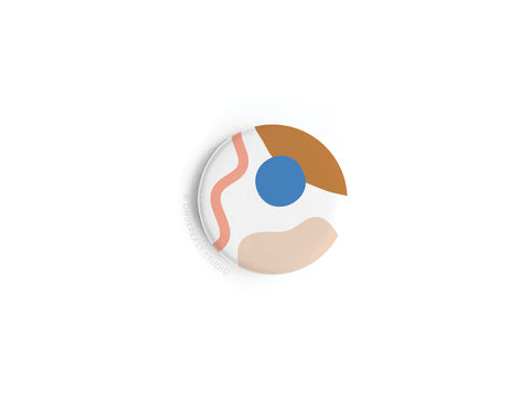 The Abstract No. 2 Button Magnet by Onderkast Studio, now discontinued, showcases a vibrant abstract design—blue circle, curved pink line, and tan and brown shapes—on white, displayed on a plain white surface.