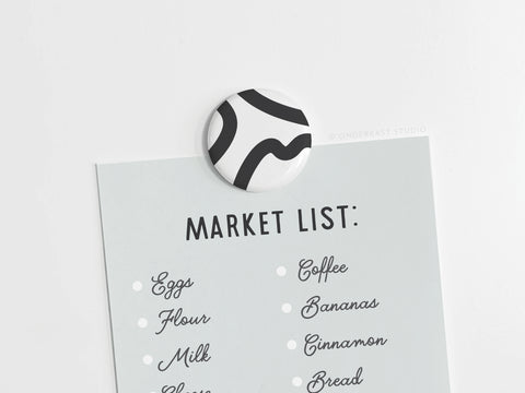 A round Abstract No. 3 Button Magnet by Onderkast Studio (DISCONTINUED), made in the USA, holds a handwritten-style Market List with items such as eggs, flour, milk, and bananas on light gray paper.