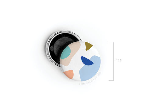 The Abstract No. 4 Button Magnet by Onderkast Studio features a colorful blue, peach, teal, olive, and beige pattern. Measuring 1.25, this discontinued magnet is printed in the USA and shown next to its black-backed open pin.