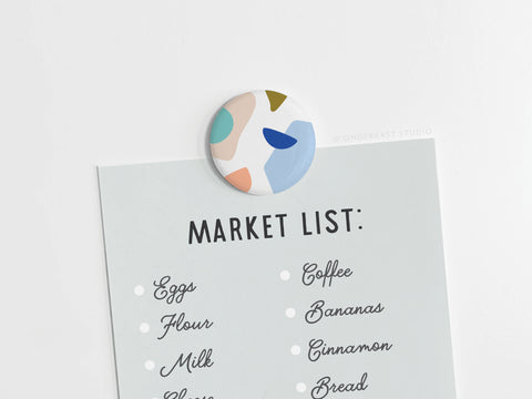 The Onderkast Studio Abstract No. 4 Button Magnet – DISCONTINUED (1.25” round, printed in the USA) holds a light blue Market List showing eggs, flour, milk, coffee, bananas, cinnamon, and bread—each marked with a white dot.