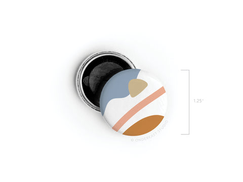 The Abstract No. 6 Button Magnet by Onderkast Studio features a vibrant blue, white, tan, orange, and peach abstract design with a diagonal stripe. The round magnet is 1.25 inches wide and partially open to show the back. Discontinued item.
