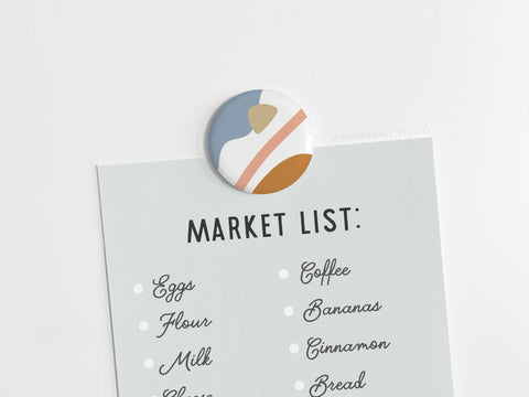An Onderkast Studio Abstract No. 6 Button Magnet (DISCONTINUED) holds a light blue “Market List:” note with a handwritten grocery list—eggs, flour, milk, coffee, bananas, cinnamon, bread—and features vibrant printed colors for quality.