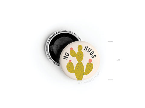 The No Hugs Cactus Button Magnet by Onderkast Studio features a green cactus with pink flowers and NO HUGS above it. At 1.25 inches, its perfect for introverts and comes with an open black-back case.