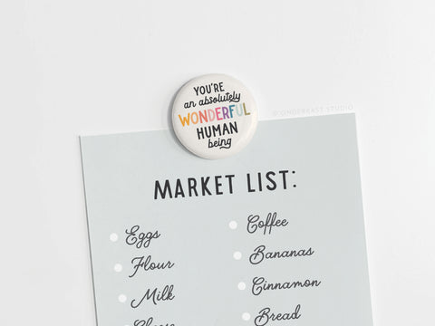 An Onderkast Studio Absolutely Wonderful Human Being Button Magnet, round and vibrantly printed in the USA, is clipped to a grocery list with eggs, flour, milk, cheese, coffee, bananas, cinnamon, and bread.
