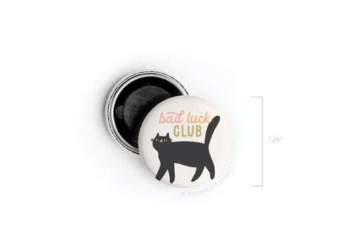 The Bad Luck Club Button Magnet by Onderkast Studio features a 1.25-inch round design with a black cat illustration and pink and yellow text, shown with its back cover slightly open for a fun, humorous touch.