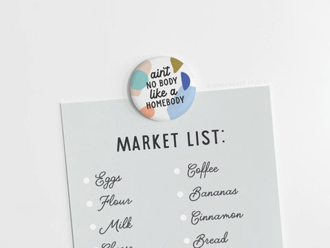 Onderkast Studio’s round, colorful Ain’t No Body Like a Homebody Button Magnet displays the phrase “aint no body like a homebody” while holding a light blue market list—eggs, flour, milk, coffee, bananas, cinnamon, bread—on a white surface.