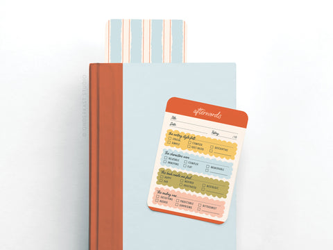 The Afterwords Book Review Card Set by Onderkast Studio sits atop a light blue and orange book, showcasing its premium cardstock, vibrant print, handwritten-style text, and sections for title, rating, themes, notes, and checkboxes.