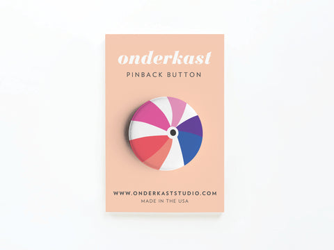 The Beachball Pinback Button by Onderkast Studio features a vibrant beach ball design on a round button, attached to a peach-colored card reading “onderkast PINBACK BUTTON” and “www.onderkaststudio.com MADE IN THE USA.”.
