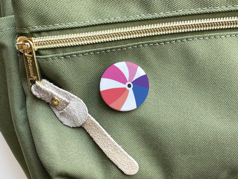 An Onderkast Studio Beachball Pinback Button with a vibrant beach ball design is pinned to a light green backpack near a gold zipper featuring a tan suede pull.