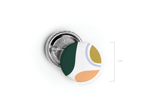 The Abstract No. 1 Pinback Button from Onderkast Studio features a vibrant green, mustard yellow, orange, gray, and white abstract design. Measuring 1.25 inches in diameter, this discontinued item is shown against a white background.