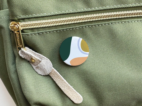 The Abstract No. 1 Pinback Button by Onderkast Studio, featuring vibrant green, white, mustard yellow, and brown colors, decorates the front pocket of a green canvas bag with a gold zipper and beige suede pull.