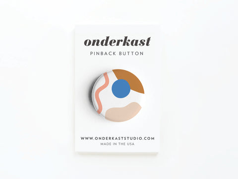 The Abstract No. 2 Pinback Button – DISCONTINUED by Onderkast Studio features abstract brown, blue, pink, and beige shapes and comes attached to a card labeled onderkast PINBACK BUTTON with website and MADE IN THE USA below.