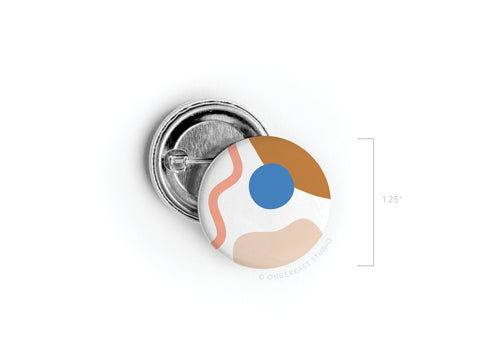 The Abstract No. 2 Pinback Button by Onderkast Studio features vibrant abstract shapes—a blue circle, tan and peach forms, and a wavy pink line—set on white. This discontinued, 1.25-inch round button is a high-quality product.