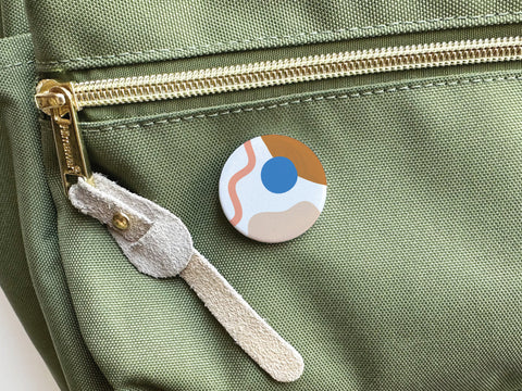 The Onderkast Studio Abstract No. 2 Pinback Button – DISCONTINUED, featuring abstract shapes and a blue dot, is shown attached to a light green fabric bag with a gold zipper and beige suede pull.