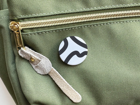 The Onderkast Studio Abstract No. 3 Pinback Button (DISCONTINUED) displays crisp black wavy lines on white, shown attached to a green fabric bag with a gold zipper and beige suede pull. Made in the USA.