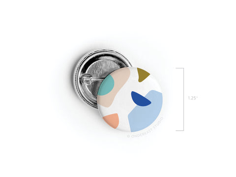The Abstract No. 4 Pinback Button by Onderkast Studio features vibrant blue, teal, peach, and olive abstract shapes on a white background. This discontinued 1.25-inch button is shown front and back with a sturdy metal pin for durability.