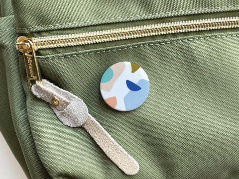 The Onderkast Studio Abstract No. 4 Pinback Button – DISCONTINUED, featuring a vibrant abstract pattern, is attached to a light green backpack near a gold zipper with a suede pull.