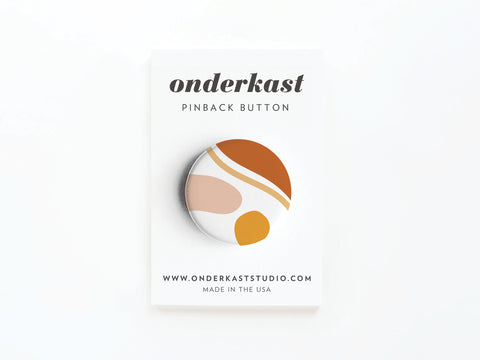 The Abstract No. 5 Pinback Button from Onderkast Studio features an abstract design in orange, gold, beige, and white with vibrant colors. Attached to a branded card with website and MADE IN THE USA text. DISCONTINUED.
