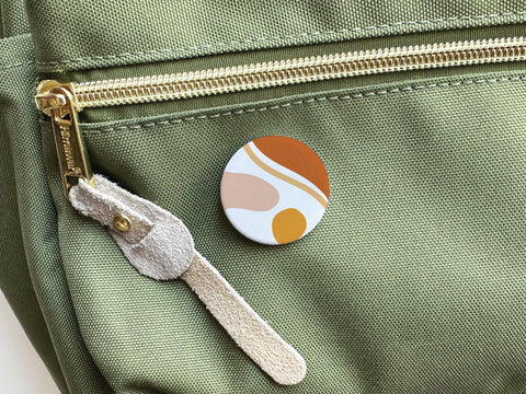 The Abstract No. 5 Pinback Button by Onderkast Studio, featuring colorful abstract shapes, is attached to a light green fabric bag with a gold zipper and beige suede pull tab.