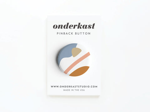 The Abstract No. 6 Pinback Button by Onderkast Studio features vibrant blue, tan, peach, and white abstract shapes on a round button, displayed on a card labeled “onderkast PINBACK BUTTON,” with website and MADE IN THE USA text below.
