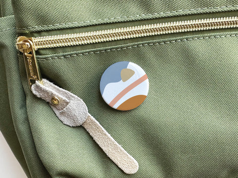 The Onderkast Studio Abstract No. 6 Pinback Button (DISCONTINUED), with a vibrant abstract landscape design, is pinned to a light green fabric bag featuring a gold zipper and beige suede zipper pull.