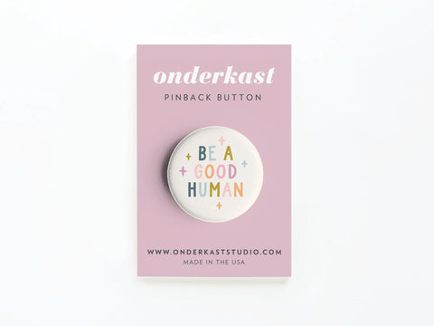 The Be a Good Human Pinback Button from Onderkast Studio features colorful lettering and comes on a pink card with the brand name and onderkaststudio.com, spreading positivity wherever you go.