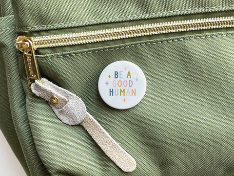 The Onderkast Studio Be a Good Human Pinback Button, with its colorful message, spreads positivity as it decorates a light green fabric bag with a gold zipper and suede pull.