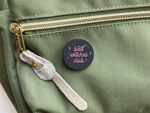 The Bad Witches Club Pinback Button by Onderkast Studio features pink lettering on black and is shown attached to a green bag with gold zipper and beige suede pull, perfect for showing off your Mystical Sisterhood style.