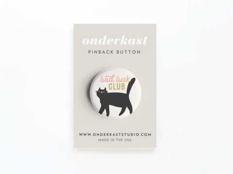 The Bad Luck Club Pinback Button by Onderkast Studio features a black cat and “Bad Luck Club” text, displayed on a card reading onderkast PINBACK BUTTON, www.onderkaststudio.com, and MADE IN THE USA.