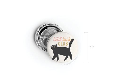 The Onderkast Studio Bad Luck Club Pinback Button is a 1.25-inch accessory with a black cat design and red/yellow text, ideal for anyone who loves playful, humorous flair.