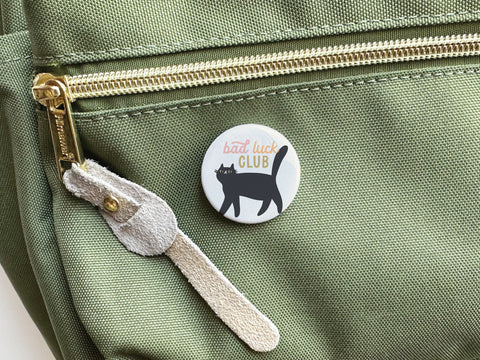 The Onderkast Studio Bad Luck Club Pinback Button is a humorous round accessory featuring a black cat and bold text, shown attached to the front pocket of a light green bag with a gold zipper and beige suede pull.
