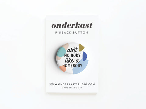 The Ain’t No Body Like a Homebody Pinback Button by Onderkast Studio features colorful abstract shapes and playful text—an ideal cozy nights accessory for introverts, displayed on a white branded card.