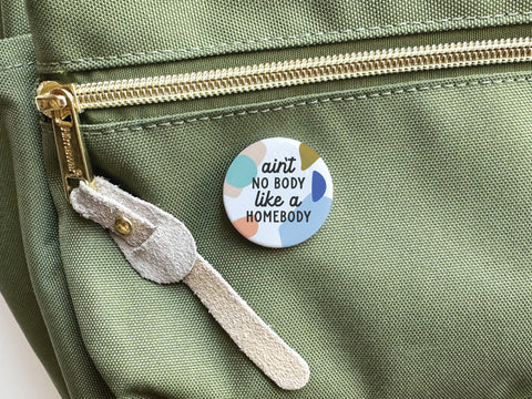 The Onderkast Studio Ain’t No Body Like a Homebody Pinback Button features colorful abstract shapes and catchy text, shown attached to a green bag with a gold zipper—a perfect accessory for homebodies.