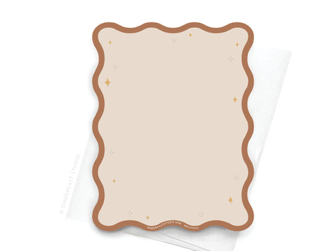 The TS Evermore Wavy Die Cut Notecard by Onderkast Studio features a blank beige card with a wavy brown border and small sparkle accents, elegantly placed on a folded white napkin.