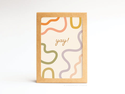 The Yay Squiggles Folded Greeting Note Set of 10 by Onderkast Studio features “yay!” in cursive on a white card with abstract peach, green, and lavender pastel squiggles, framed in brown. Printed in the USA.