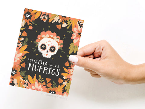 A hand holds the Feliz Día de los Muertos Greeting Card – FINAL STOCK by Onderkast Studio, featuring colorful floral designs and a decorated skull—perfect for celebrating tradition and remembrance on the Day of the Dead.