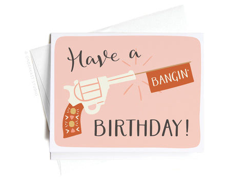 The Have a Bangin’ Birthday! Greeting Card by Onderkast Studio features a cartoon revolver with a patterned handle shooting a BANGIN flag, plus the message Have a BANGIN BIRTHDAY! on a pink background.