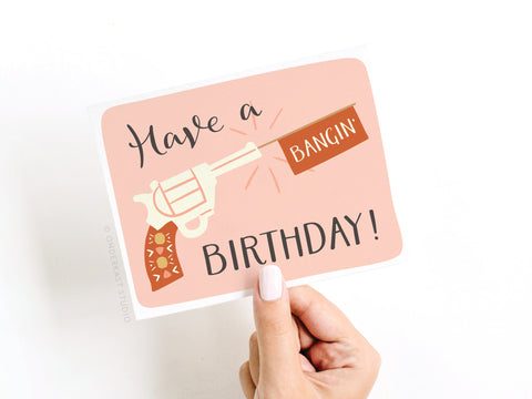 A hand holds the Have a Bangin’ Birthday! Greeting Card by Onderkast Studio, featuring playful fonts and a toy gun shooting a BANGIN flag on a lively pink background.