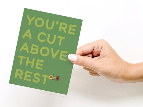 A hand with white nails holds the Youre a Cut Above the Rest Lawn Mower Greeting Card by Onderkast Studio, featuring a lawnmower cutting through the word REST and the phrase YOURE A CUT ABOVE THE REST.