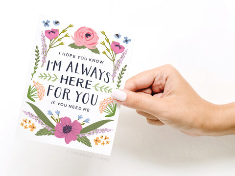 A hand holds the Onderkast Studio I’m Always Here For You Greeting Card, adorned with pink, purple, and yellow flowers plus green leaves. Printed in the USA, this card is a heartfelt reminder of support.