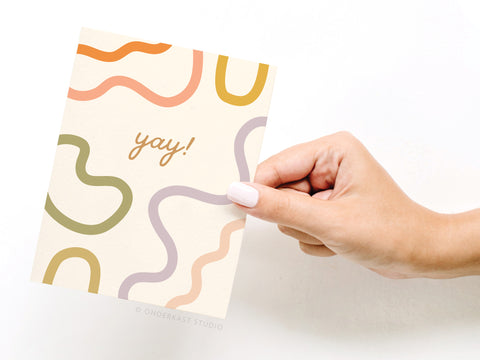 A hand with light pink nail polish holds the Yay Squiggles Greeting Card by Onderkast Studio, featuring colorful abstract squiggles and yay! in cursive on a cream background.
