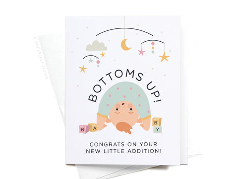 The Bottoms Up Baby Greeting Card by Onderkast Studio features a playful upside-down baby with a star and moon mobile. Text: BOTTOMS UP! Congrats on your new little addition! Perfect for congratulating new parents.